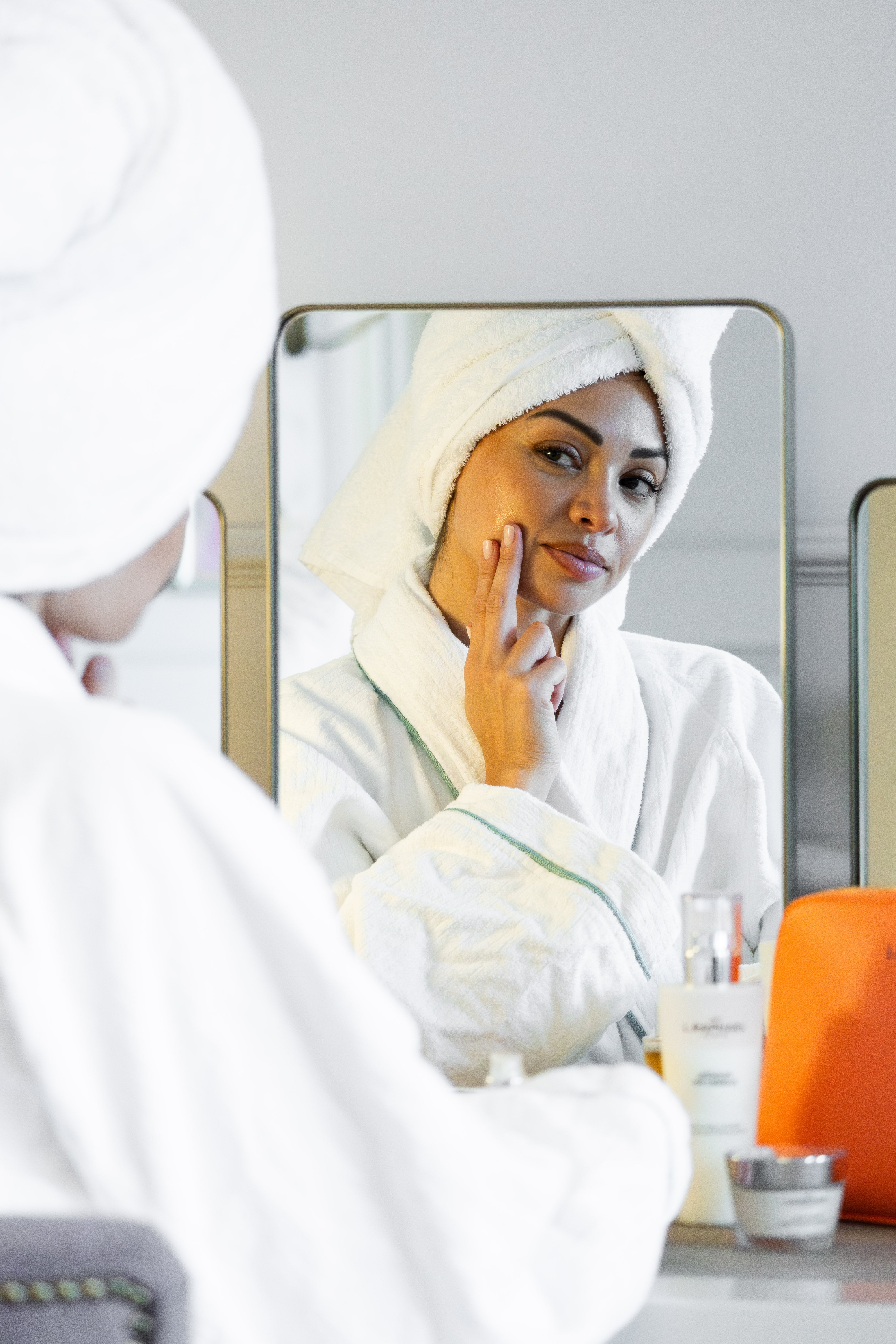 Woman applying skincare product in front of a mirror, wearing a towel on her head.