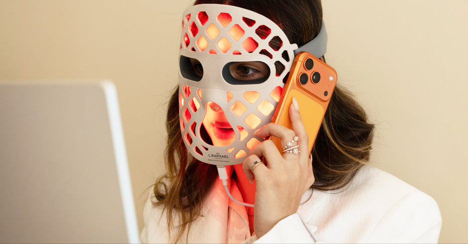 A woman wearing a white LED facial therapy mask with orange light shining through, holding an orange smartphone to her ear while looking at a computer screen.