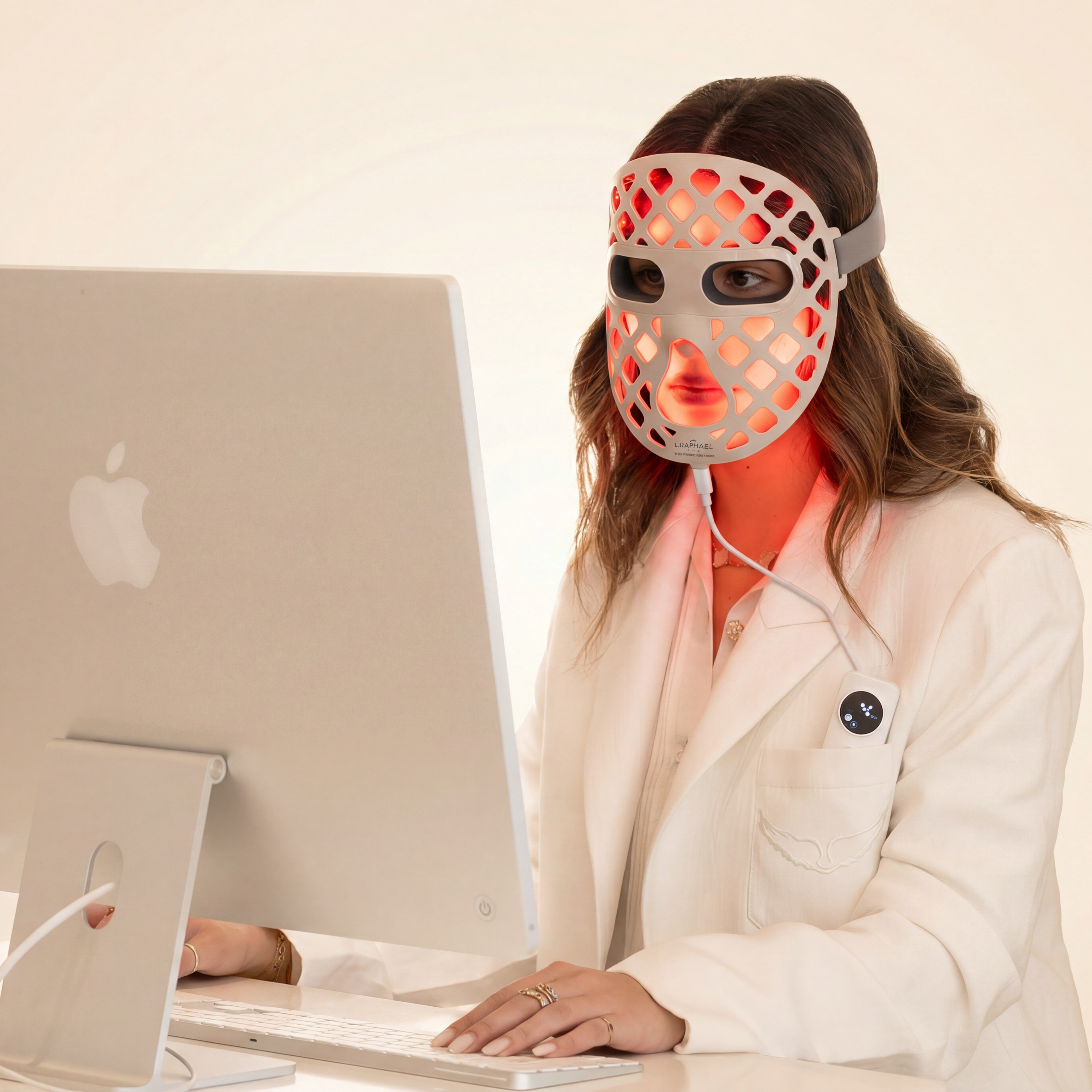 A woman in a white lab coat sits at a desk using a computer, wearing a red and orange LED light therapy mask on her face.